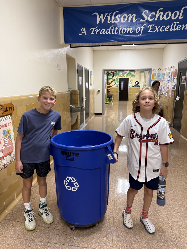 2 students, standing in hallway, holding recycle bin