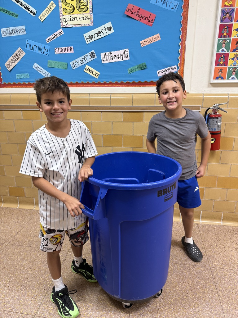 2 students, standing in the hallway, with the recycling bin