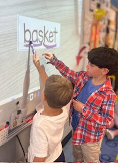 two first grade students, standing at the board, scooping the word basket