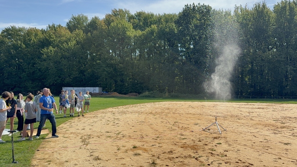 A group of students and adults stand on a grassy field and dirt baseball diamond, watching a water bottle rocket launch into the air, creating a cloud of water mist above its launcher.