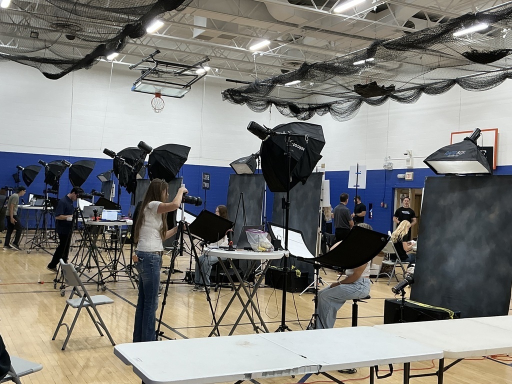 A gymnasium set up with multiple photography stations for picture day, complete with backdrops, studio lighting strobes, softboxes, and cameras on tripods. Photographers are assisting students who are seated or standing for their portraits.