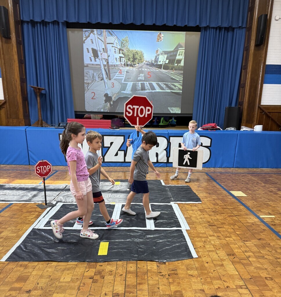 Elementary students practice pedestrian safety inside a gymnasium on a simulated crosswalk mat with street lane lines. One student holds a stop sign and another holds a pedestrian crossing sign while classmates walk across. A projected image on a screen behind them shows a real intersection.