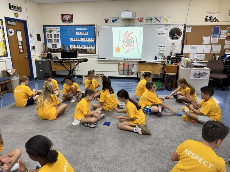Elementary students wearing yellow shirts that read "RESPECT" on the back sit in a circle on a classroom rug during a music lesson, participating in a rhythmic activity.