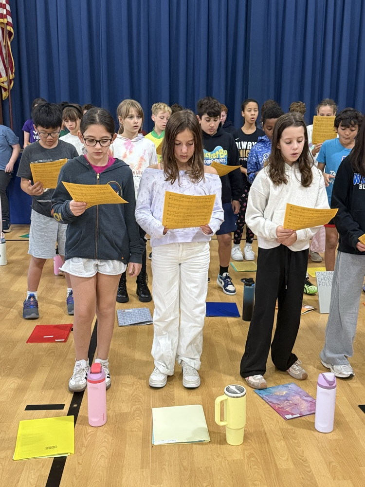 An elementary school choir standing in rows on a wooden stage, holding yellow sheet music and singing during a chorus rehearsal in front of a blue backdrop curtain.