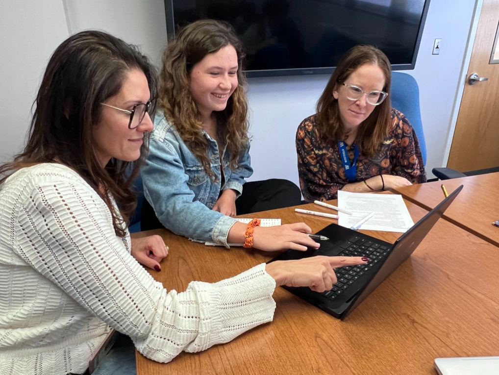 A student and two educators sit together at a wooden conference table, looking at a laptop screen. The adult on the left points at the laptop screen while discussing information with the student and staff member beside her.