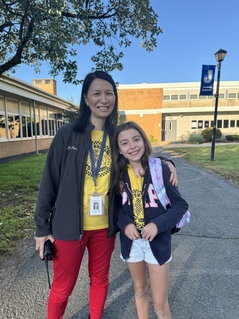 The principal standing outdoors alongside a young girl wearing a yellow shirt and dark zip-up hoodie in front of Jefferson School.