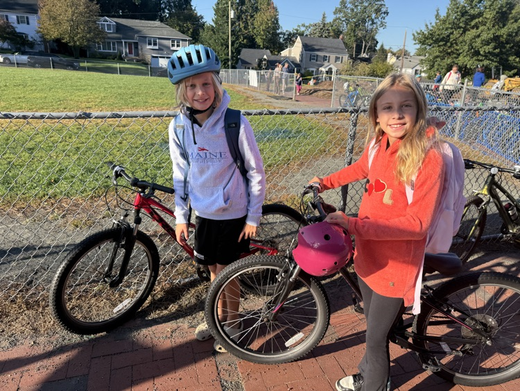 Two young students standing with their bicycles on a brick path next to a chain-link fence, both holding or wearing bike helmets.