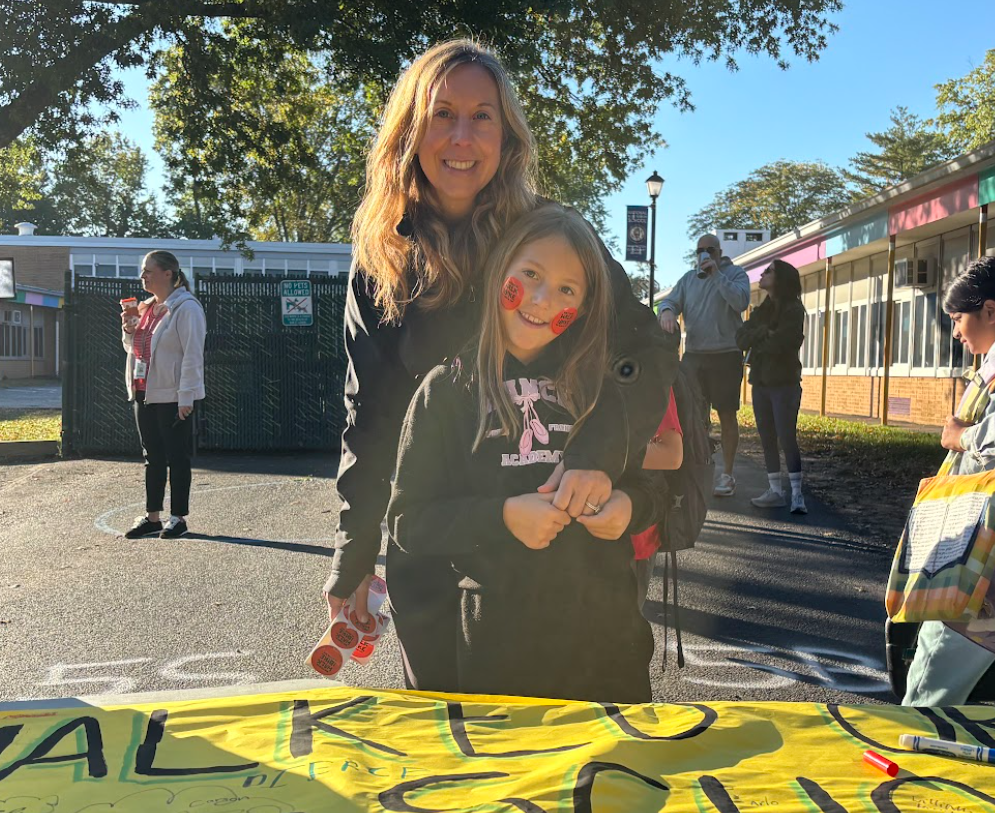 A smiling woman standing outdoors next to a young girl wearing stickers on her cheeks in front of a yellow banner that reads "Walk to School."