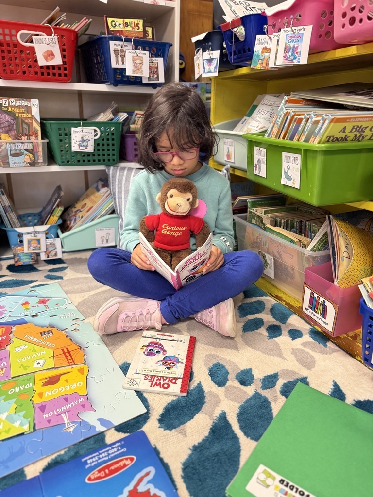 A young student wearing pink glasses and a green sweater sits cross-legged on a patterned rug in a classroom reading corner, holding a small stuffed monkey wearing a red "Curious George" shirt while reading a book. Educational games, puzzles, and colorful book bins fill the surrounding shelves.   
