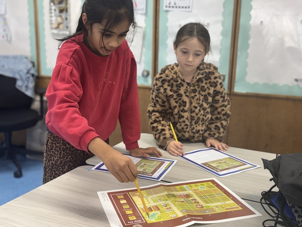 Two elementary students stand at a table leaning over printed farm maps, using pencils to trace paths and locate key map elements using a key at the bottom.   