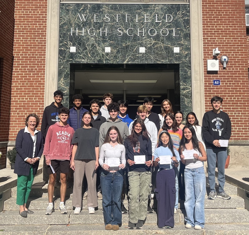 A group of Westfield High School students stands on the outdoor front steps of the school building beneath the stone "Westfield High School" entrance sign. Several students hold paper certificate awards, accompanied by the principal
