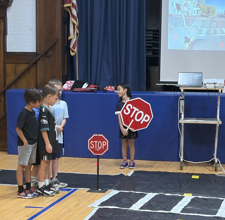 Elementary students practice pedestrian safety inside a gym set up with a simulated road and crosswalk mat. One student stands holding a large octagon "STOP" sign while three classmates stand lined up behind a small mounted stop sign waiting to cross.   