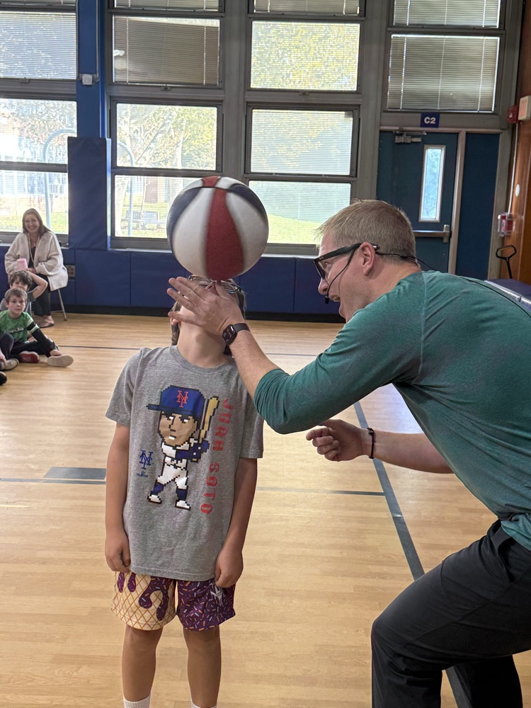 In a gymnasium setting, a presenter wearing glasses and a green long-sleeved shirt balances a red, white, and blue basketball on a young student's extended fingers. The student wears a gray shirt depicting a baseball player.   