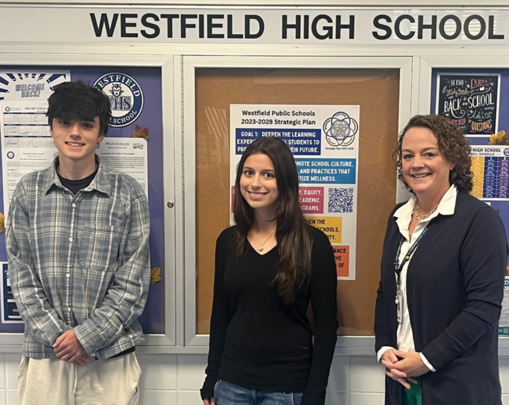 Students Alexander Stepanov and Emma Simpson stand alongside Westfield High School Principal Asfendis in a school hallway in front of a glass bulletin board labeled "WESTFIELD HIGH SCHOOL". 