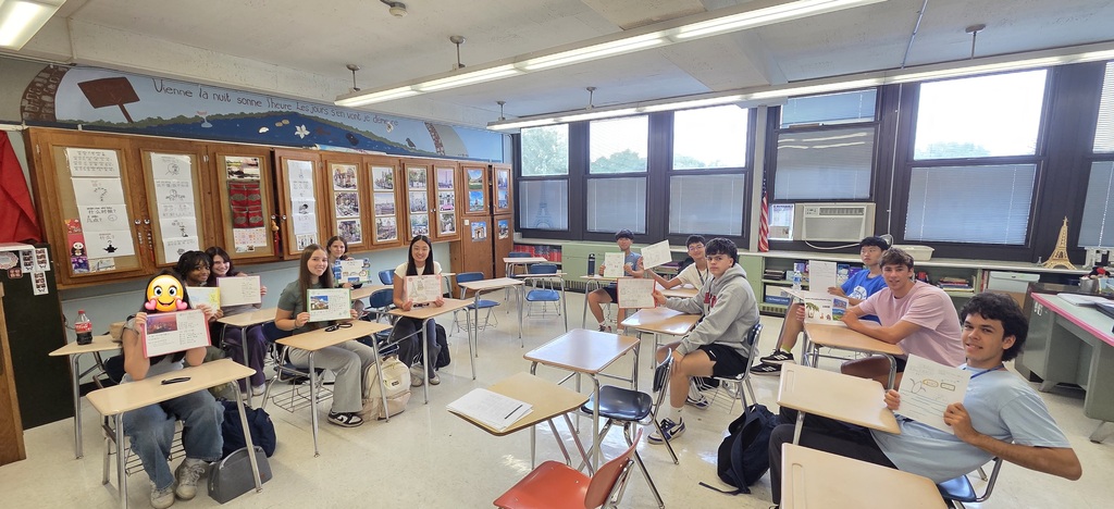 Students sit at individual desks arranged in a high school language classroom, holding up hand-written and hand-drawn paper travel advertisements. The classroom features large windows along the back wall, displays of student work on the cabinets, and a painted mural above the bulletin board.