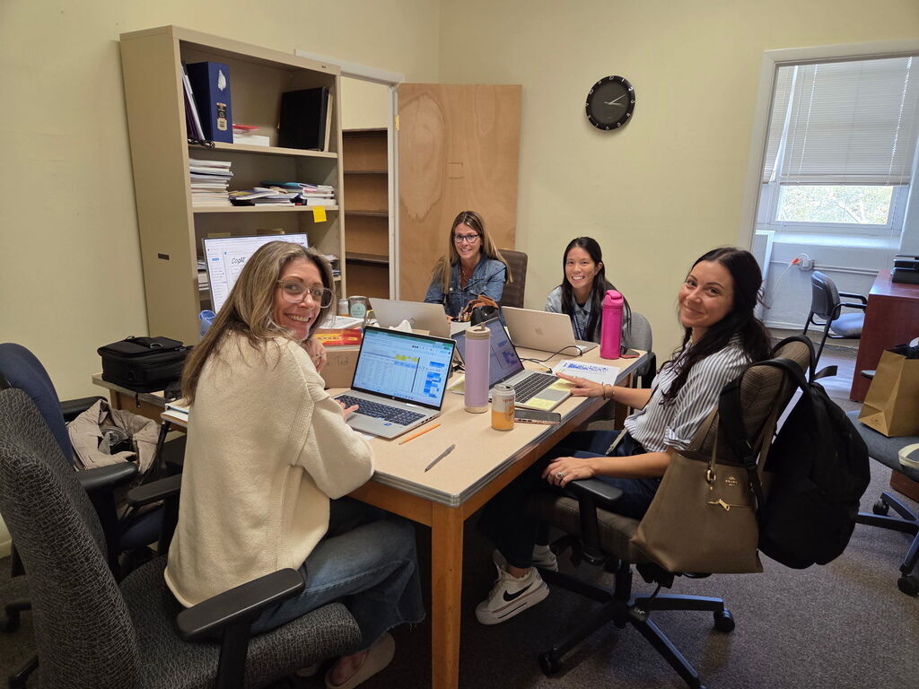 Four educators smiling at the camera while seated around a wooden desk with laptops, papers, and office supplies in a school workspace.   