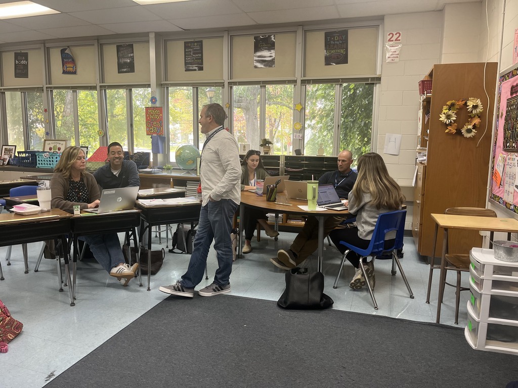 A group of teachers sitting at desks arranged in a circle in a classroom, listening and engaged as a male colleague stands to speak to the group.   