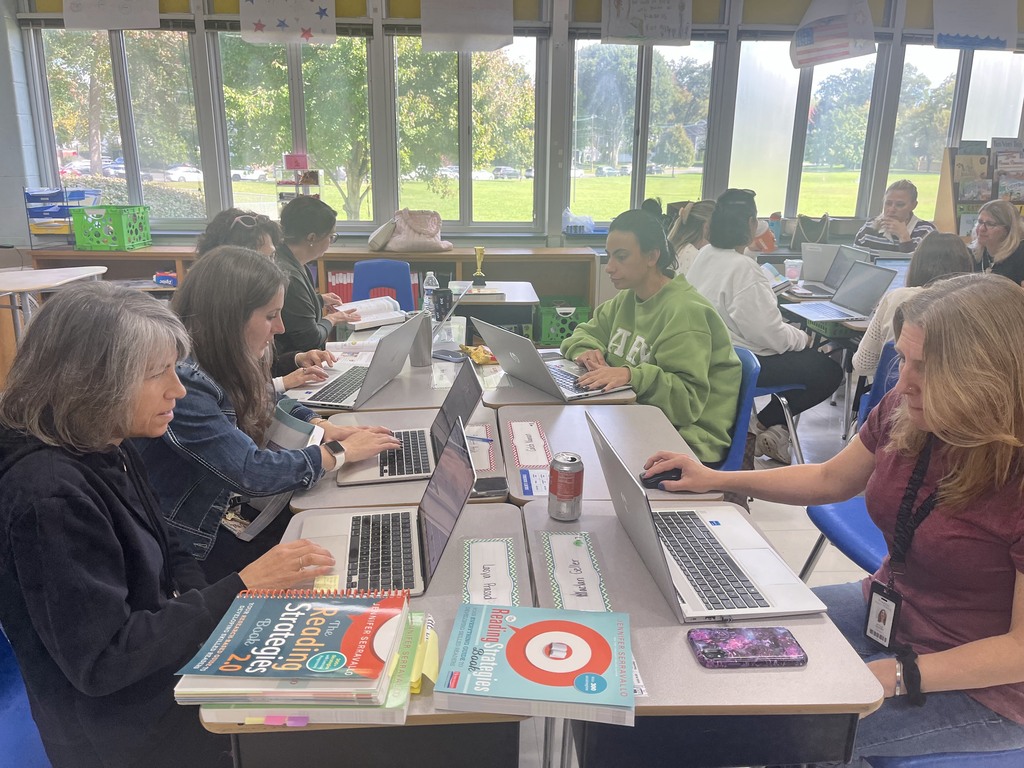 A close-up view of teachers sitting around connected desks in a classroom, actively typing on laptops with professional reading strategy books open in front of them.   