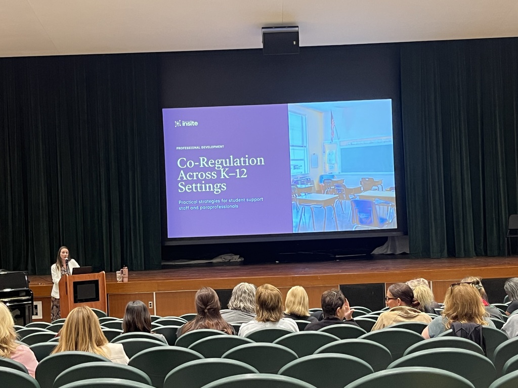 An auditorium presentation showing a large screen with a slide titled "Co-Regulation Across K–12 Settings" while a presenter stands at the podium addressing educators seated in the rows ahead.   