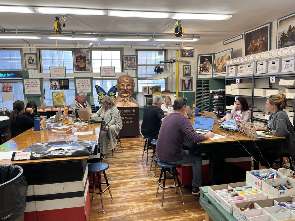 Teachers seated around high wooden tables in a spacious art room equipped with art supplies and historical posters, working collaboratively on their laptops.   