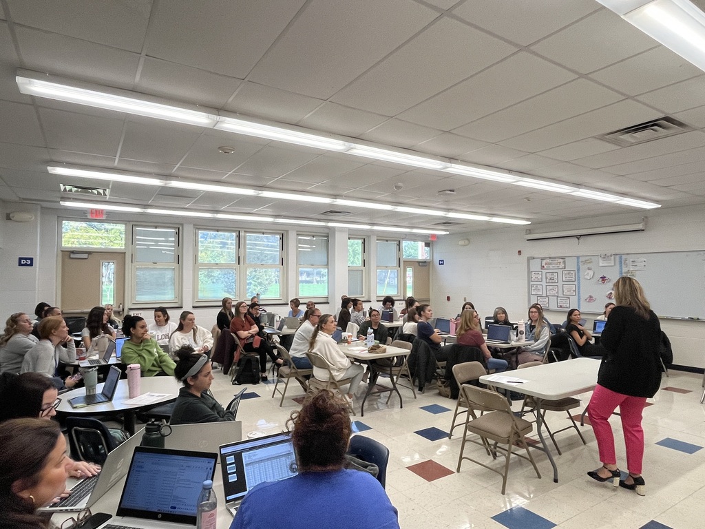 A presenter in a black jacket and pink pants speaks to a large group of teachers seated at tables with laptops inside a bright classroom.   