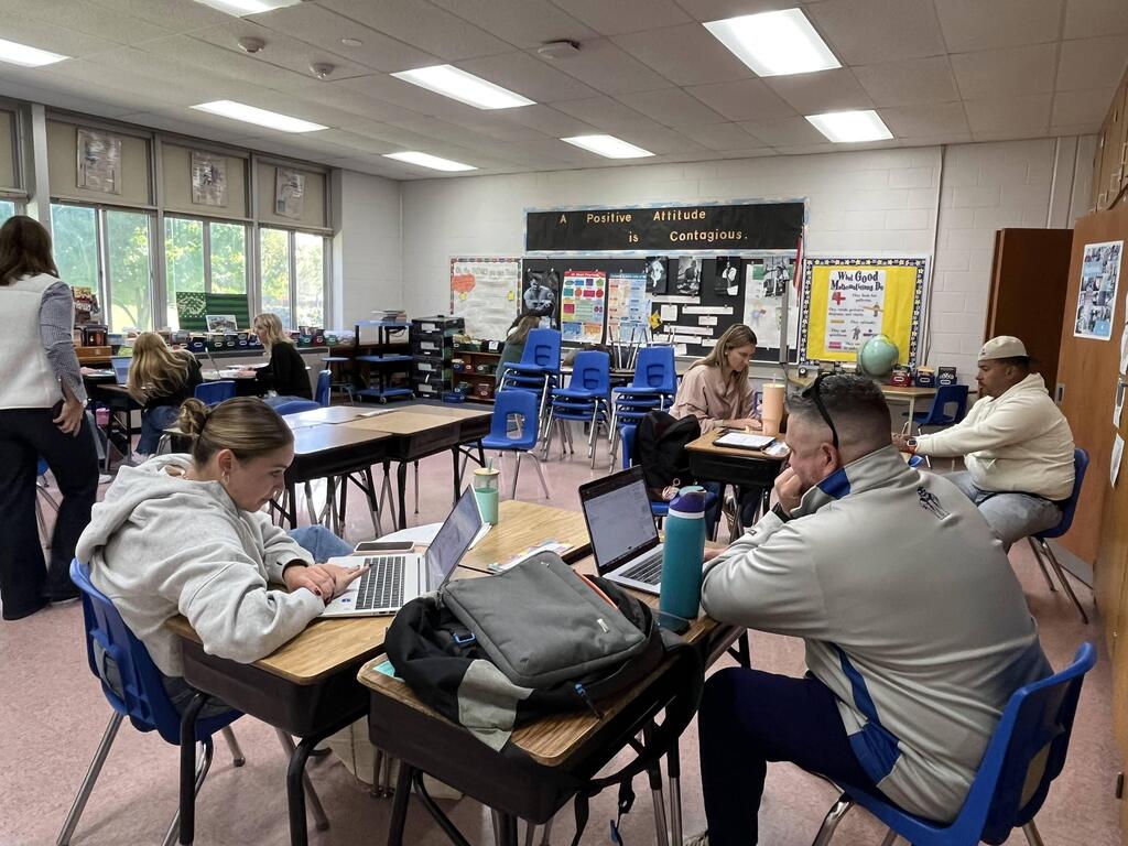 Teachers sitting at individual student desks arranged in small discussion clusters inside a classroom, working on laptops under a bulletin board reading "A Positive Attitude is Contagious."   