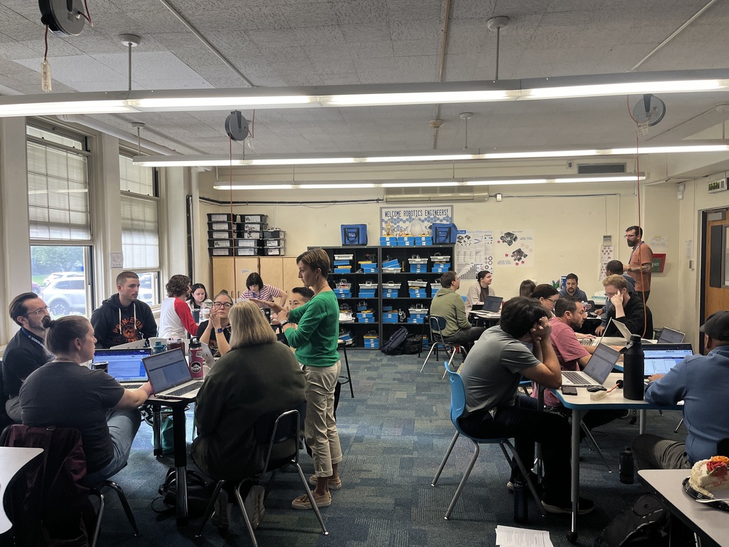 A group of teachers working together in a robotics and STEM classroom, seated around tables with laptops while an instructor in a green top facilitates the discussion.   