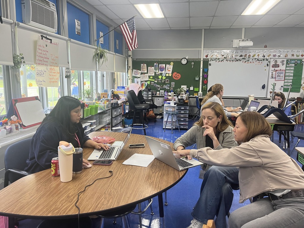 Three educators sitting at a curved wooden table in a decorated elementary classroom, looking closely at a laptop screen together to collaborate on lesson planning.   
