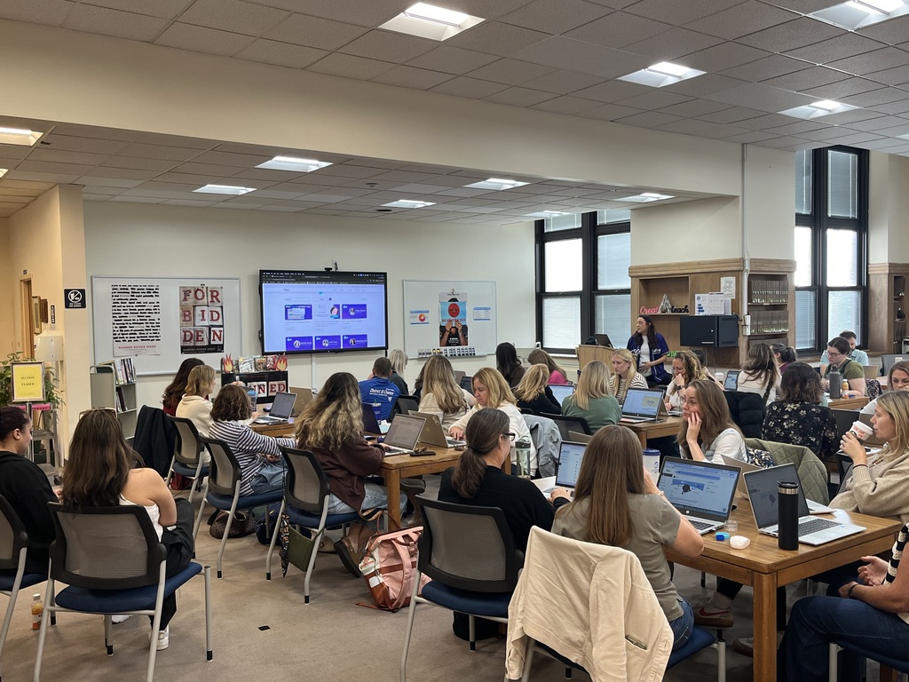 A wide shot of educators seated at large wooden tables inside a school library media center, working together on laptops during a professional development session with a digital screen and posters displayed at the front.   
