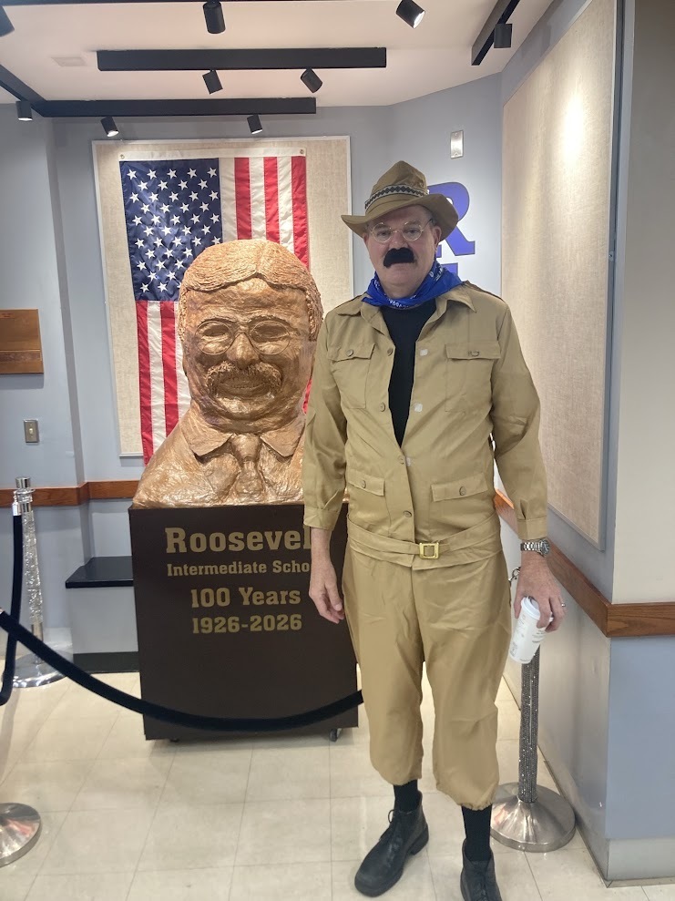 An adult dressed in a khaki safari outfit, round glasses, and a fake mustache stands next to a large bronze bust of Theodore Roosevelt in front of an American flag.
