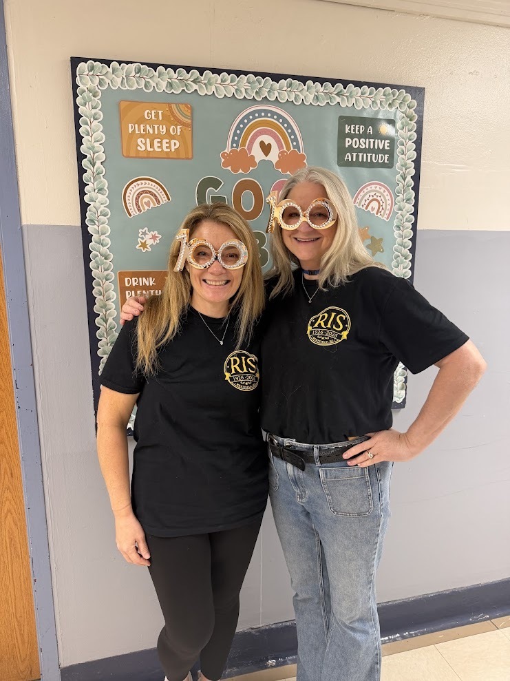 Two adults stand in front of a bulletin board decorated with rainbows and the message Get Plenty of Sleep, both wearing festive 100-year celebration glasses and matching RIS black t-shirts.