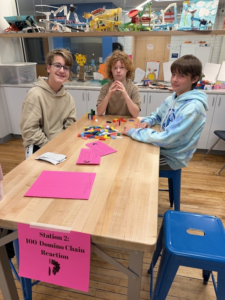 Three students sit at a wooden table examining colorful dominoes and pink cards labeled Station 2