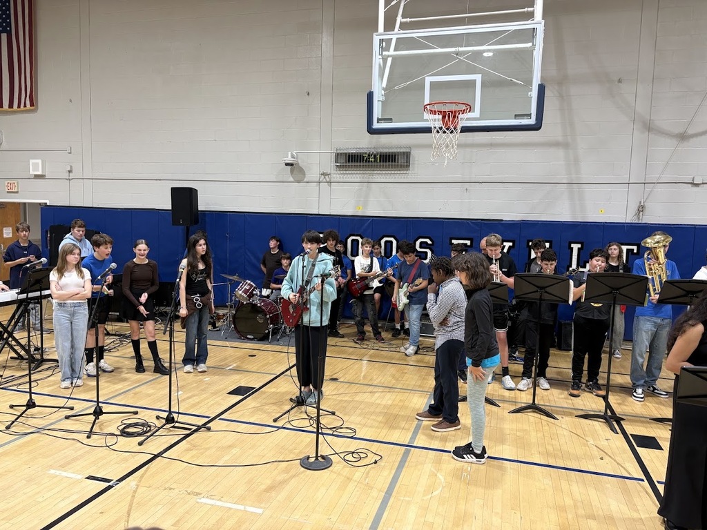 Students perform with instruments and vocals on a gym floor in front of a blue padded wall, with a basketball hoop overhead.