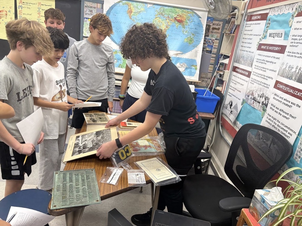 Students gather around a wooden table looking at historical photos and a framed plaque in a classroom with a world map on the wall.
