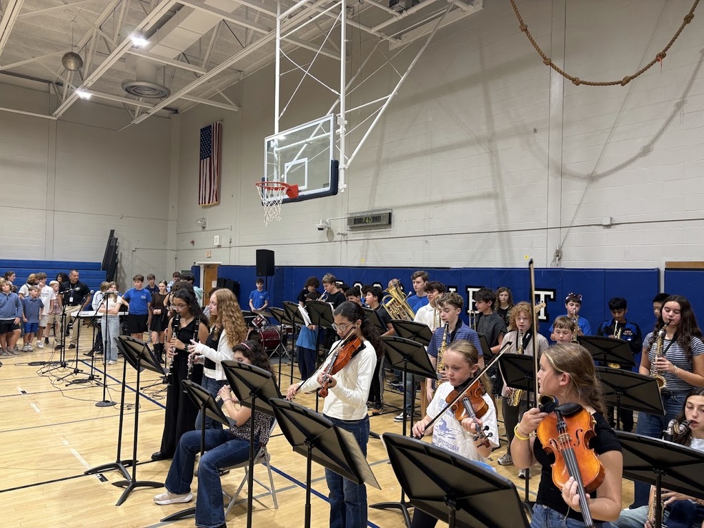 Students play string and wind instruments, reading from music stands in a gymnasium.
