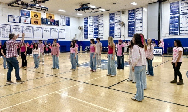 A student vocal group dressed in pink and casual attire standing in a semi-circle on the gym floor performing under the direction of an instructor during the assembly.