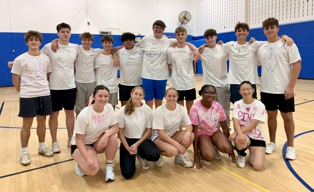 A group of male and female students in pink and white shirts posing together for a photo on the basketball court inside the gymnasium.
