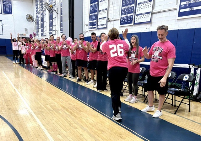 A group of Westfield High School staff members wearing matching bright pink shirts with the number 26 high-fiving and celebrating on the gymnasium court during the volleyball event.