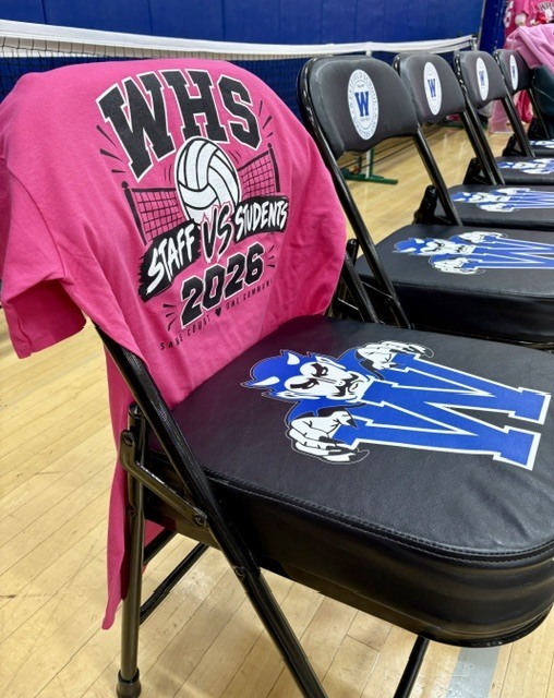 A close-up shot of a bright pink t-shirt displaying the text "WHS Staff vs Students 2026" draped over the back of a black folding sideline chair bearing the Westfield Blue Devils mascot logo.