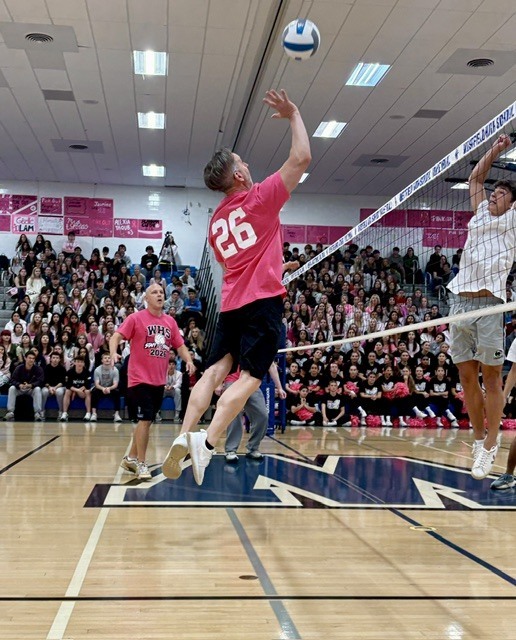 An action shot during a volleyball match in a crowded gymnasium, showing a staff member in a pink shirt jumping near the net to spike the ball while a student on the opposing team jumps to block it.