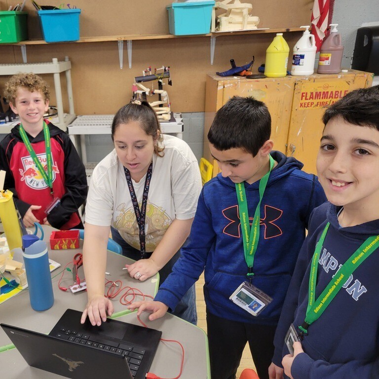 A teacher wearing green lanyards sits at a table guiding two middle school students wearing green Edison Intermediate School lanyards as they work with wires connected to a laptop.