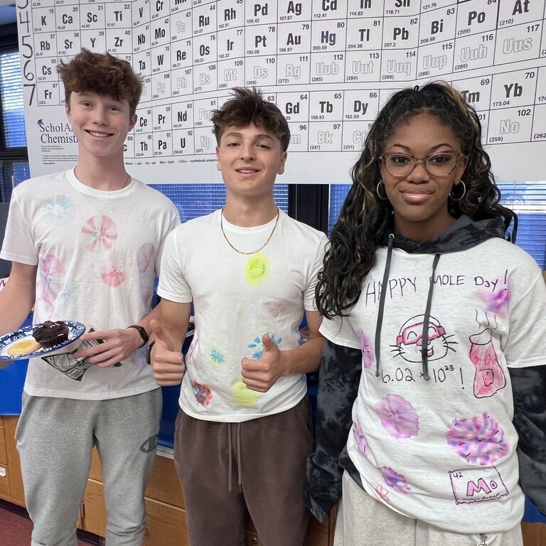 Three high school students pose together in front of a periodic table poster, with one student wearing a tie-dye shirt that reads "Happy Mole Day!" and features chemistry formulas.
