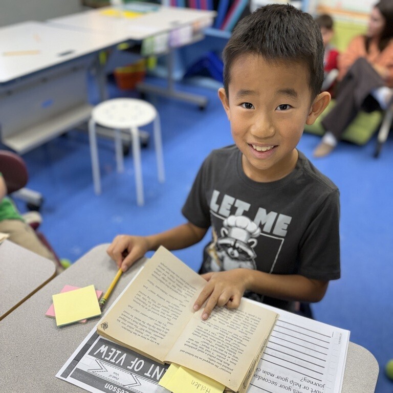 An elementary student smiles at the desk while holding open a book next to a worksheet titled "Point of View" and yellow sticky notes.