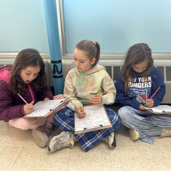 Three elementary students sit cross-legged on a tiled classroom floor in front of a blue pole, holding clipboards and writing with pencils.