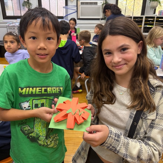 A younger student and an older student hold up a 3D orange paper craft craft project together in a classroom setting.