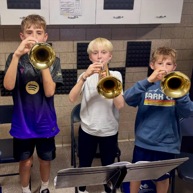 Three middle school students stand side by side holding brass wind instruments up to their mouths in a music practice room.