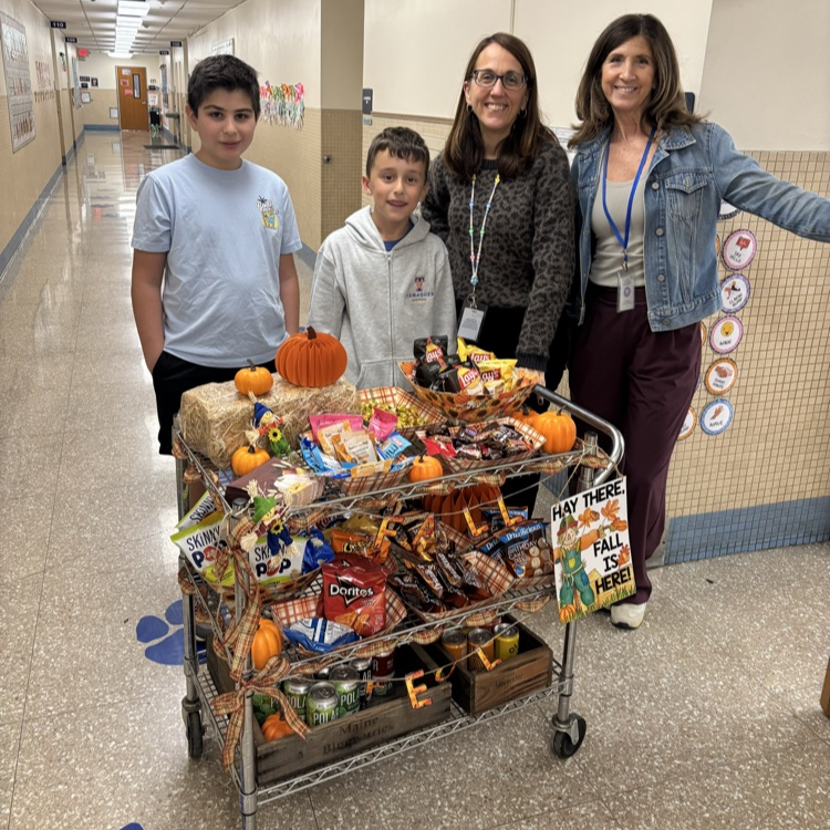 Two staff members stand beside two students in a school hallway next to a rolling cart filled with fall-themed snacks, treats, and decorative pumpkins.