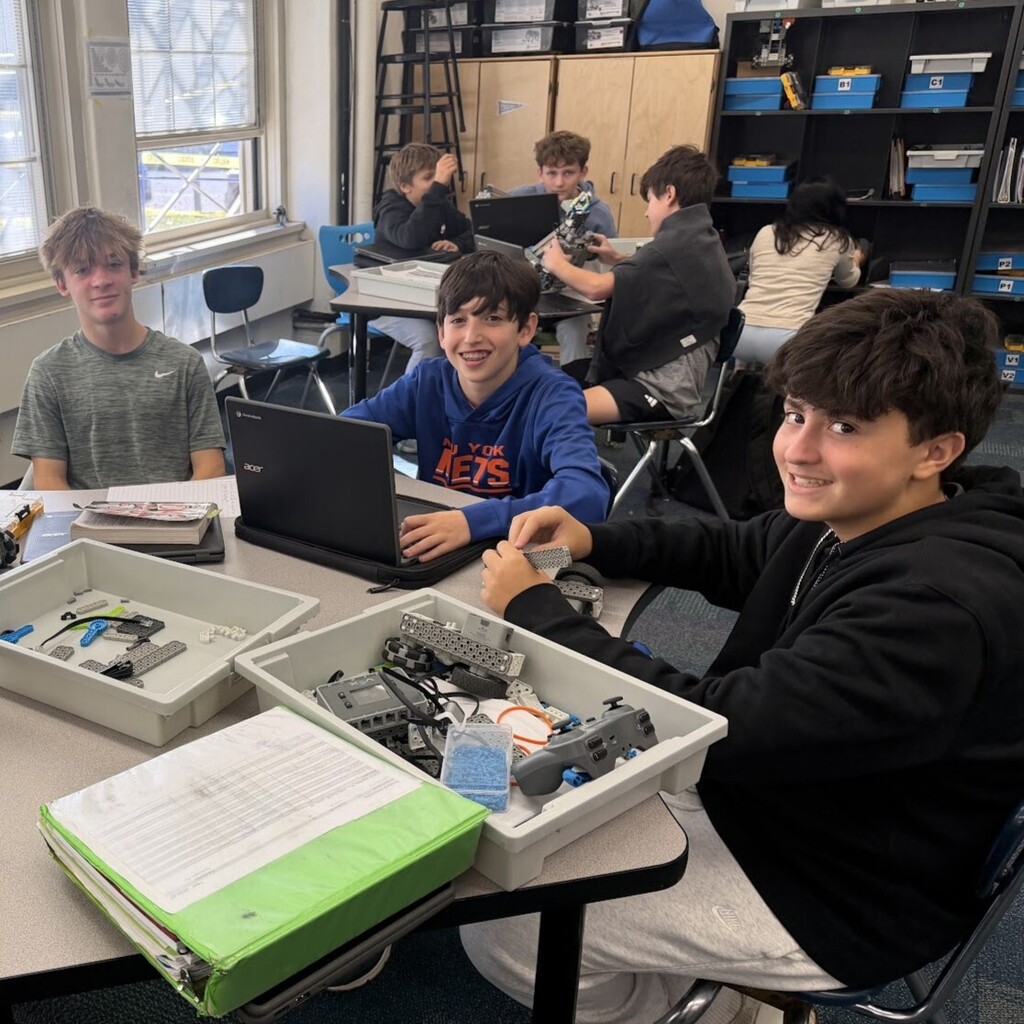 Three students sit together around a desk in a STEM classroom with open laptop computers, plastic storage tubs filled with robotics parts, and a game controller.