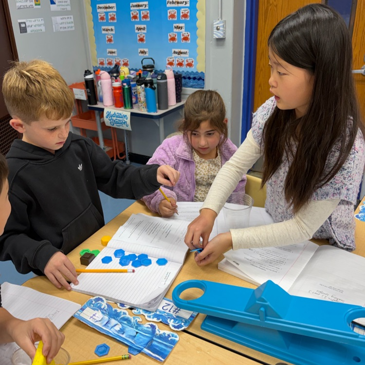 Three elementary students collaborate around a table using blue hexagonal tiles and pencils with open notebooks during a classroom lesson.
