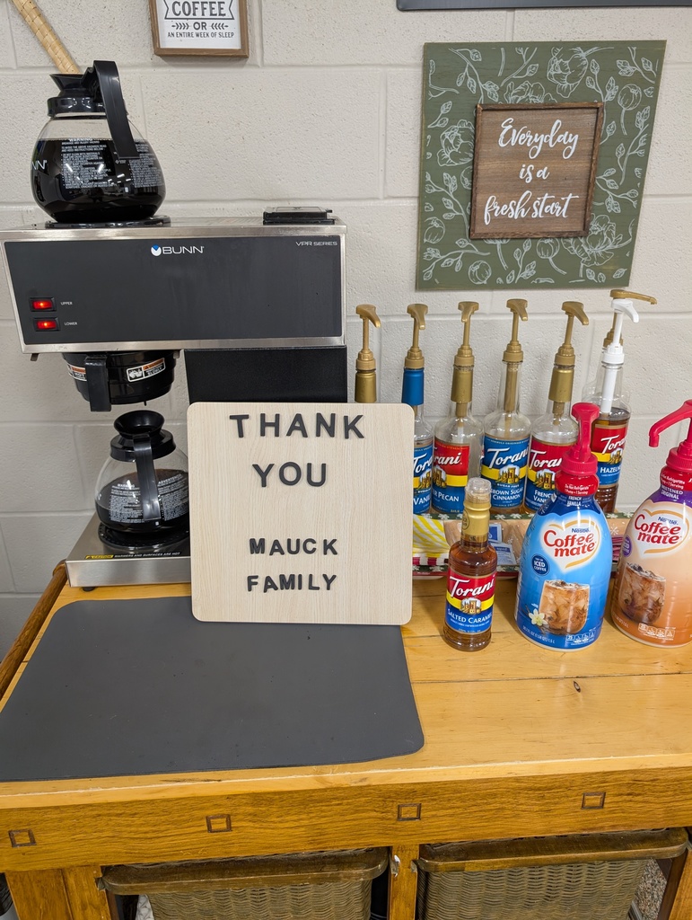 "A well-stocked coffee station on a wooden counter. The station includes a professional BUNN coffee brewer with two glass carafes, several bottles of Torani syrups (Vanilla, Butter Pecan, Brown Sugar Cinnamon, and French Vanilla), and Coffee-mate creamers. A central wooden letter board reads 'THANK YOU MAUCK FAMILY.' In the background, a decorative green sign says 'Everyday is a fresh start.'"